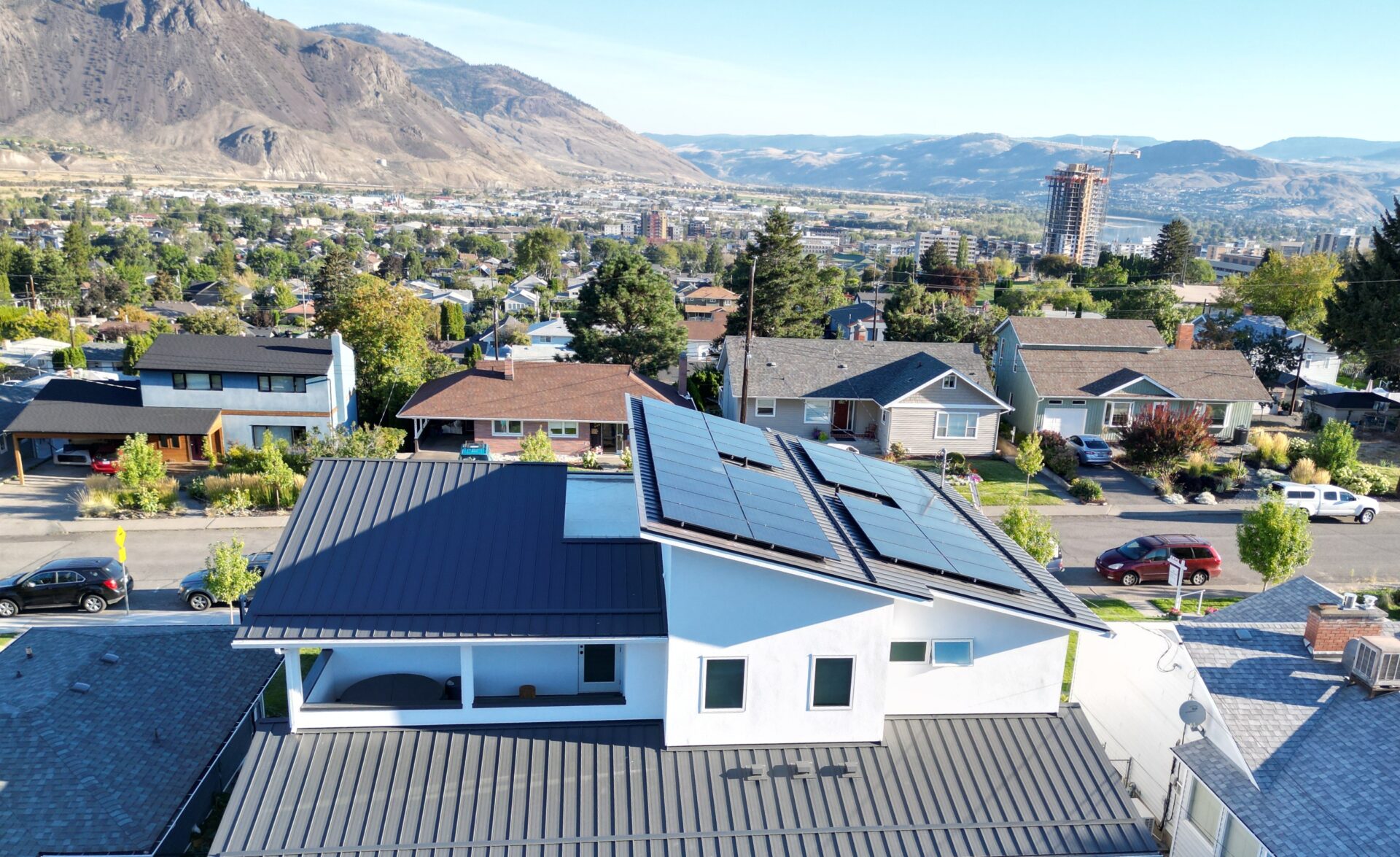 An aerial view shows a modern house with solar panels installed on a dark metal roof in a quiet suburban neighbourhood. Surrounding homes, tree-lined streets, and parked cars create a residential setting, while a tall building under construction rises in the distance. Rolling mountains and a broad valley form a scenic backdrop under a clear sky.
